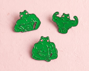 Könnte beinhalten: Drei grüne Emaille-Anstecknadeln mit muskulösen Froschfiguren mit rosa Wangen. Jede Anstecknadel zeigt eine andere Pose eines Frosches mit definierter Bauch- und Armmuskulatur, dargestellt im Cartoon-Stil mit schwarzen Umrandungen.