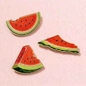 May include: Three enamel pins shaped like watermelon slices. Each pin features a bright red fruit interior with black seeds, a thin white rind, and a green outer edge, all outlined in gold metal. The slices are depicted in different orientations.