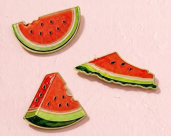 Könnte beinhalten: Drei Emaille-Anstecknadeln in Form von Wassermelonenscheiben. Jede Nadel zeigt ein leuchtend rotes Fruchtfleisch mit schwarzen Kernen, eine dünne weiße Schale und einen grünen Außenrand, alles eingefasst in goldfarbenes Metall.