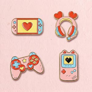 Könnte beinhalten: Ein Set aus vier Emaille-Anstecknadeln mit Gaming-Motiven: eine Handheld-Konsole mit rotem Herz-Bildschirm, ein rosa Kopfhörer mit herzförmigen Ohrmuscheln, ein rosa Gamecontroller mit roten Tasten und eine vertikale Konsole mit Pixel-Herz.