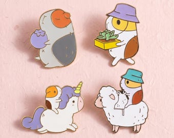 Könnte beinhalten: Vier Emaille-Anstecknadeln mit Meerschweinchen-Motiven. Eines hält eine violette Beere, eines trägt einen violetten Hut und eine Sukkulente, eines reitet auf einem weißen Einhorn mit violetter Mähne und eines reitet auf einem weißen Schaf.