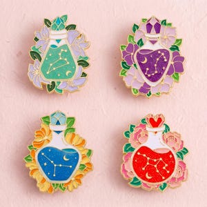 Può includere: Quattro spille in smalto a forma di boccette di pozione, ognuna contenente un liquido di colore diverso con una costellazione dorata e una luna crescente. Le boccette sono circondate da fiori colorati nelle tonalità viola, giallo e rosa.