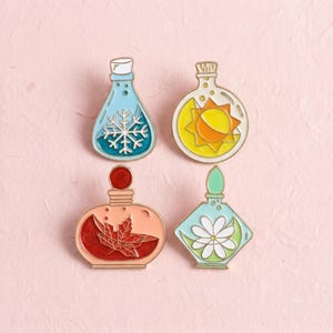 Könnte beinhalten: Vier Emaille-Anstecknadeln in Form von Glasflaschen, die jeweils ein anderes saisonales Symbol enthalten: eine weiße Schneeflocke in einer blauen Flasche, eine gelbe Sonne, ein rotes Ahornblatt in einer pfirsichfarbenen Flasche und eine weiße Blume.