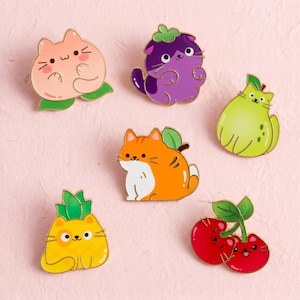 May include: A collection of six enamel pins featuring cat-themed fruit designs. The set includes a peach cat, a purple eggplant cat, a green pear cat, an orange tabby cat, a yellow pineapple cat, and a pair of red cherry cats with green leaves.