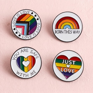 Könnte beinhalten: Vier runde Emaille-Anstecknadeln mit Pride-Motiven. Eine zeigt eine Progress-Pride-Flagge mit dem Text LOVE IS LOVE. Eine weitere zeigt einen Regenbogenbogen mit BORN THIS WAY. Eine dritte zeigt ein Regenbogenherz mit YOU ARE SAFE WITH ME. Die letzte zeigt ein Regenbogenherz mit JUST LOVE.