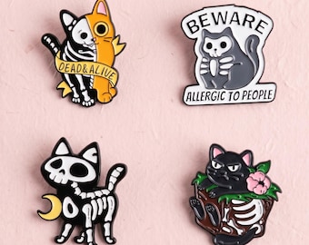 Könnte beinhalten: Vier Emaille-Anstecknadeln mit Katzenmotiven. Eine zeigt eine halb-Skelett-Katze mit dem Text DEAD & ALIVE. Eine weitere ist eine graue Katze mit Totenkopfgesicht und dem Text BEWARE ALLERGIC TO PEOPLE. Eine dritte ist eine schwarze Skelett-Katze mit gelbem Mond. Eine vierte ist eine schwarze Katze in einer Kiste mit Skelett und einer rosa Blume.