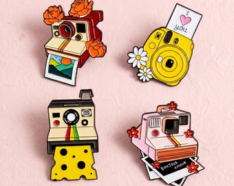 Könnte beinhalten: Vier Emaille-Anstecknadeln in Form von Vintage-Kameras. Eine ist rot mit orangefarbenen Blumen, eine gelb mit weißen Gänseblümchen und einem 'I love you'-Foto, eine schwarz mit Käsemuster und eine rosa mit roten Blumen verziert.