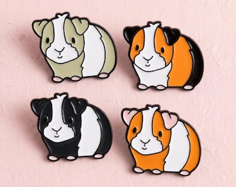Könnte beinhalten: Vier Emaille-Anstecknadeln in Form von Meerschweinchen. Eine Nadel ist beige-weiß, eine orange-weiß-schwarz, eine schwarz-weiß und eine orange-weiß mit hellrosa Ohren. Jede Anstecknadel zeigt ein Tierdesign im Cartoon-Stil.