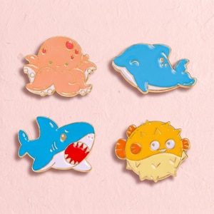 May include: A set of four enamel pins featuring sea creatures: an orange octopus, a blue dolphin, a blue shark with an open mouth showing white teeth, and a yellow pufferfish with orange fins. Each pin has a gold-toned metal outline.