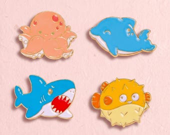 Könnte beinhalten: Ein Set aus vier Emaille-Anstecknadeln mit Meeresbewohnern: ein orangefarbener Oktopus, ein blauer Delfin, ein blauer Hai mit offenem Maul und weißen Zähnen sowie ein gelber Kugelfisch mit orangefarbenen Flossen. Jede Nadel hat einen Goldrand.