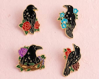 Könnte beinhalten: Vier Emaille-Anstecknadeln mit schwarzen Raben und goldfarbenen Umrandungen. Jeder Rabe ist mit kleinen goldenen Sternen und Mondsicheln verziert. Die Vögel sind von bunten Blumenakzenten umgeben, darunter rote Rosen, blaue und violette Blüten.