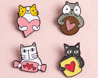 Könnte beinhalten: Vier Emaille-Anstecknadeln mit Cartoon-Katzen. Eine ist eine weiß-gelbe Katze, die ein rosa Herz hält. Eine andere ist eine graue Katze, die einen braunen Kreis mit einem rosa Herz hält. Eine weiße Katze hält ein rosa Rechteck mit drei roten Herzen. Eine schwarze Katze hält ein gelbes Quadrat mit einem roten Herz.