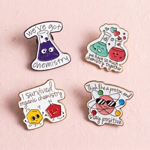 Könnte beinhalten: Vier Emaille-Anstecknadeln mit Chemie-Motiven. Eine zeigt einen lila Kolben mit dem Text 'we've got chemistry'. Eine andere zeigt einen grünen und einen roten Kolben mit Herzen und 'we have so much chemistry together'. Eine dritte zeigt ein gelbes Sechseck und ein rotes Fünfeck mit 'I survived organic chemistry'. Die letzte zeigt ein Atom mit Sonnenbrille und 'Think like a proton and stay positive'.