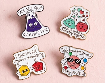 Könnte beinhalten: Vier Emaille-Anstecknadeln mit Chemie-Motiven. Eine zeigt einen lila Kolben mit dem Text 'we've got chemistry'. Eine andere zeigt einen grünen und einen roten Kolben mit Herzen und 'we have so much chemistry together'. Eine dritte zeigt ein gelbes Sechseck und ein rotes Fünfeck mit 'I survived organic chemistry'. Die letzte zeigt ein Atom mit Sonnenbrille und 'Think like a proton and stay positive'.