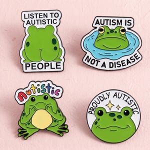 Könnte beinhalten: Vier Emaille-Anstecknadeln mit grünen Fröschen und Text. Die Aufschriften lauten LISTEN TO AUTISTIC PEOPLE, AUTISM IS NOT A DISEASE, AUTISTIC in Regenbogenfarben und PROUDLY AUTISTIC mit zwei kleinen gelben Sternen über dem Froschkopf.