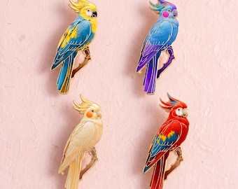 Könnte beinhalten: Vier Emaille-Anstecknadeln in Form von Kakadus, die auf Ästen sitzen. Die Nadel oben links ist gelb und blau, oben rechts blau und violett, unten links cremefarben und unten rechts rot, blau und gelb, jeweils mit goldenen Umrandungen versehen.