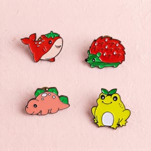 Könnte beinhalten: Vier Emaille-Anstecknadeln mit Tiermotiven: ein roter Erdbeerwal, ein roter Erdbeerigel, ein rosafarbener Erdbeerdinosaurier und ein gelber Frosch mit einem grünen Blatt auf dem Kopf. Jede Anstecknadel hat eine glatte Oberfläche und farbige Konturen.