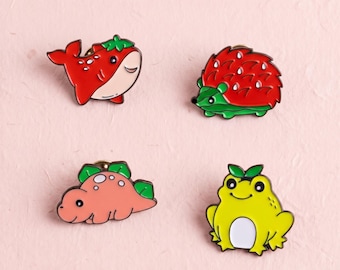 Könnte beinhalten: Vier Emaille-Anstecknadeln mit Tiermotiven: ein roter Erdbeerwal, ein roter Erdbeerigel, ein rosafarbener Erdbeerdinosaurier und ein gelber Frosch mit einem grünen Blatt auf dem Kopf. Jede Anstecknadel hat eine glatte Oberfläche und farbige Konturen.
