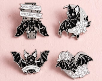 Könnte beinhalten: Vier schwarz-weiße Emaille-Anstecknadeln mit Fledermausmotiven. Eine Nadel zeigt eine Fledermaus auf einem Sarg mit dem Text VAMPIRES WILL NEVER HURT YOU. Eine weitere zeigt eine Fledermaus mit einer Mondsichel und Blumen sowie eine mit dem Text batty for you.