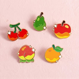 May include: A set of five enamel pins featuring fruit designs including a pair of red cherries, a green pear, a red and yellow apple, a red and yellow peach, and an orange with a green leaf, all accented with small white star-shaped highlights.