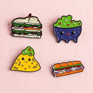 Könnte beinhalten: Ein Set aus vier Emaille-Anstecknadeln mit Lebensmittelmotiven: ein geschichtetes Sandwich, eine Schüssel mit grünem Guacamole mit lächelndem Gesicht, ein gelber Nacho-Chip mit lächelndem Gesicht und ein langes Sub-Sandwich mit Belag.