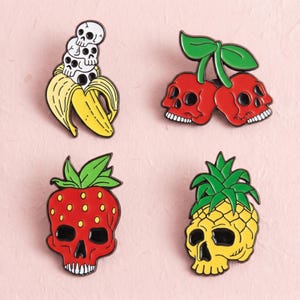 Op de afbeelding: Vier emaille pins met fruit- en schedelontwerpen. Eén pin toont een gele banaan met drie witte schedels gestapeld. Een andere toont twee rode kers-schedels met groene bladeren. Een derde is een rode aardbei-schedel en de vierde is een gele ananas-schedel.