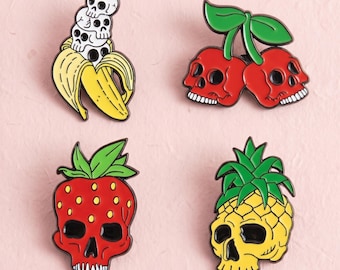Könnte beinhalten: Vier Emaille-Anstecknadeln mit Frucht- und Totenkopfmotiven. Eine Nadel zeigt eine gelbe Banane mit drei gestapelten weißen Totenköpfen. Eine weitere zeigt zwei rote Kirsch-Totenköpfe mit grünen Blättern. Zudem gibt es einen roten Erdbeer-Totenkopf und einen gelben Ananas-Totenkopf.
