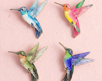 Könnte beinhalten: Vier Kolibri-Emaille-Anstecknadeln in leuchtenden Farbkombinationen. Eine ist blau-weiß, eine rosa-gelb, eine grün-orange und eine lila-grün. Jeder Vogel ist im Flug mit ausgebreiteten Flügeln und goldfarbenen Metallumrandungen dargestellt.