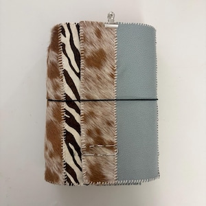 Pode incluir: Uma capa de caderno retangular com um design de retalhos que combina pele de vaca castanha e branca, uma secção com padrão de zebra a preto e branco e um painel texturizado azul claro. As secções são unidas por costuras brancas.