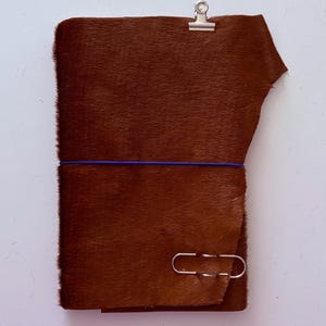May include: A rectangular notebook cover made of brown hair-on hide leather. The cover is secured with a thin, horizontal blue elastic band. A silver metal binder clip is attached to the top edge, and a silver paper clip is attached to the bottom.