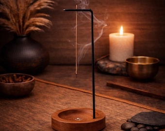May include: A wooden incense holder with a black metal arm, holding a burning incense stick. Smoke rises from the incense. A lit candle, a small brass bowl, and a decorative vase are in the background. The scene is set on a wooden surface.
