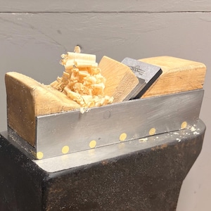 May include: A hand plane made of metal and wood, with a collection of wood shavings. The plane features a silver body, wooden components, and a metal blade. The shavings are light brown and rest on the plane.