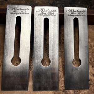 May include: Three rectangular metal plane blades with a brushed finish. Each blade features a central vertical slot ending in a circular hole. The text Roberts Fine Tools MagnaMax is engraved at the top of each individual steel component.