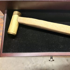 May include: A wooden mallet with a brass head rests inside a dark drawer. The mallet has a light-colored wooden handle and a cylindrical brass striking surface. The drawer is lined with black fabric and has a small, silver-colored handle.