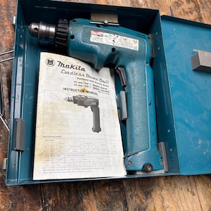 Könnte beinhalten: Ein türkisfarbener Makita Akku-Bohrschrauber in einem offenen blauen Metallkoffer, mit einer Bedienungsanleitung. Der Bohrschrauber hat ein schwarzes Bohrfutter. Das Handbuch lautet "Makita Cordless Driven Drill".