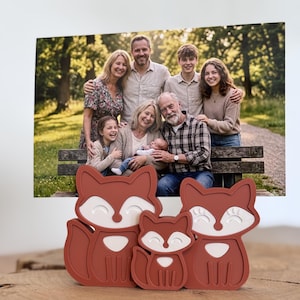 Può includere: Tre decorazioni a forma di volpe di diverse dimensioni, realizzate in materiale marrone rossiccio con dettagli bianchi. Sullo sfondo, una foto di famiglia, con un gruppo di persone che posano all'aperto. Le volpi sono posizionate davanti alla foto.