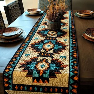 Puede incluir: Camino de mesa acolchado con diseño del suroeste. El camino tiene un fondo crema con patrones geométricos en turquesa, naranja, marrón y negro. Está sobre una mesa de madera, con platos y un jarrón con flores secas. Mide aprox. 180 cm.