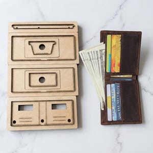 May include: A set of three light-coloured wooden leathercraft cutting dies with serrated edges alongside a brown leather bifold wallet containing several credit cards and US dollar bills tucked into the interior slots and main compartment.