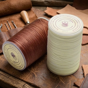 May include: Two spools of thread, one brown and one off-white, sit on a leather surface. The brown spool has a label that reads "Specializing in the manufacture of a variety of handmade..." The off-white spool has a label with the number "002".