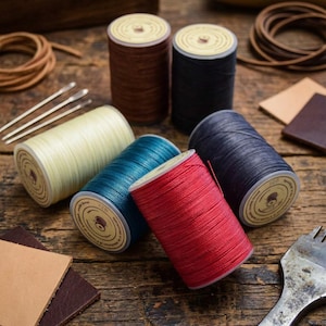 May include: A collection of colorful spools of thread in various shades, including brown, navy, teal, red, and cream. The spools are arranged on a rustic wooden surface, alongside leather scraps, needles, and other crafting tools.