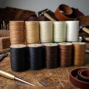 May include: A selection of waxed thread spools in shades of brown, beige, black, and gold, displayed on a wooden surface. The cylindrical spools are neatly arranged, indicating a craft or leatherworking focus. Tools and leather pieces are also visible in the background.