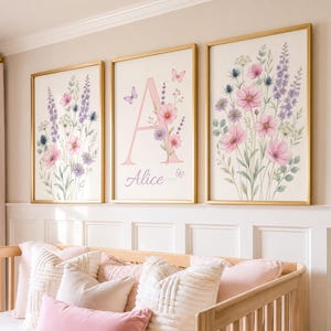 Peut inclure: Trois cadres muraux présentant des illustrations à l'aquarelle de fleurs sauvages roses et violettes. L'affiche centrale affiche une grande lettre A rose et le prénom Alice en écriture cursive, agrémentée de petits papillons et de fleurs.