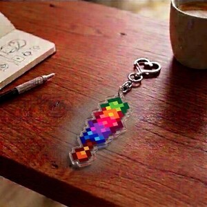 May include: A colorful pixelated sword keychain with a silver heart-shaped clasp. The sword design features a vibrant gradient of rainbow colors. The keychain is made of clear acrylic and rests on a wooden surface.