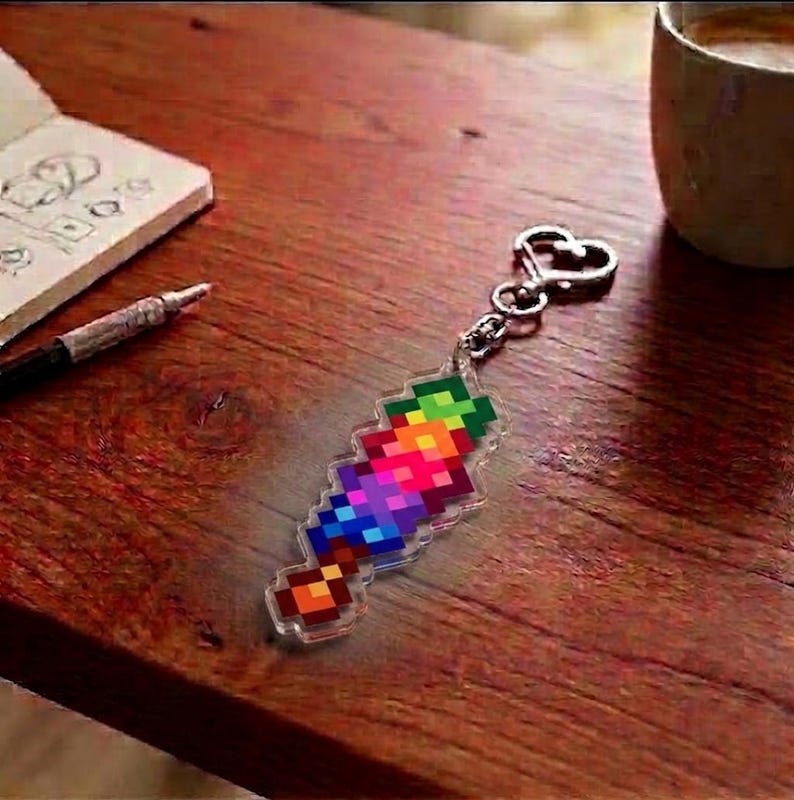 May include: A colorful pixelated sword keychain with a silver heart-shaped clasp. The sword design features a vibrant gradient of rainbow colors. The keychain is made of clear acrylic and rests on a wooden surface.