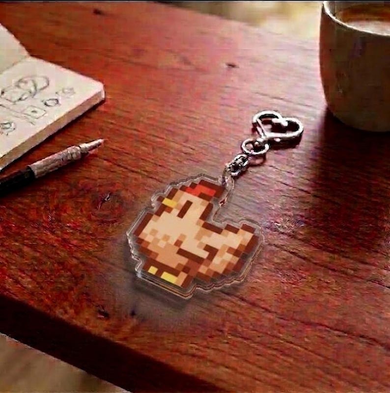 May include: A pixelated chicken keychain with a silver heart-shaped clasp. The keychain is made of clear acrylic and features a brown, orange, and yellow design. The keychain is on a wooden surface, next to a notebook, pen, and a cup.