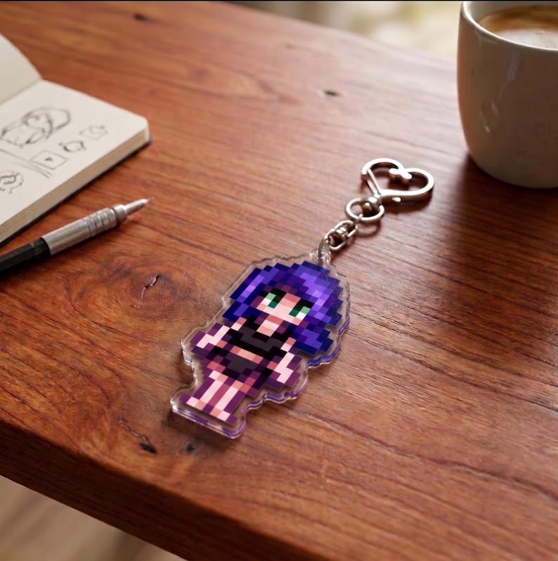 May include: A pixelated acrylic keychain featuring a female character with purple hair and green eyes. The keychain is attached to a silver heart-shaped clasp. The background includes a wooden surface, a notebook, a pen, and a coffee cup.