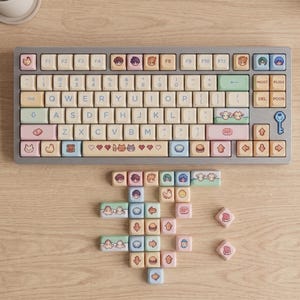 May include: A gray mechanical keyboard with pastel-colored keycaps. The keycaps feature cartoon illustrations and symbols. Some keycaps are scattered around the keyboard on a wooden surface. The keyboard has a key with a key symbol.