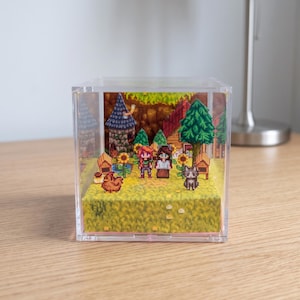 May include: A clear acrylic cube diorama featuring a pixelated farm scene. The scene includes two figures, a chicken, a cat, sunflowers, a house, a tower, and trees. The base is a yellow field.