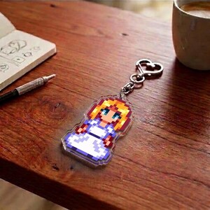 May include: A pixelated keychain featuring a character with blonde hair, blue eyes, and a white dress. The keychain is attached to a silver heart-shaped clasp. The background includes a wooden surface, a notebook, a pen, and a coffee mug.