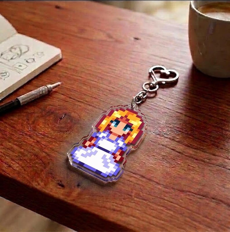 May include: A pixelated keychain featuring a character with blonde hair, blue eyes, and a white dress. The keychain is attached to a silver heart-shaped clasp. The background includes a wooden surface, a notebook, a pen, and a coffee mug.